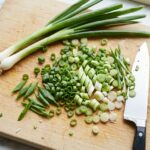 A chef's knife rests on a light wood cutting board alongside a pile of freshly chopped scallions and two whole stalks.