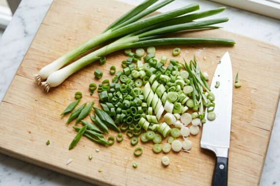 A chef's knife rests on a light wood cutting board alongside a pile of freshly chopped scallions and two whole stalks.