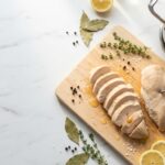 An overhead shot of a wooden cutting board placed on a white marble countertop, featuring sliced and whole pieces of cooked chicken breast.