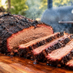 Sliced smoked brisket with dark bark and a juicy interior on a wooden cutting board, with a barbecue smoker in the background.