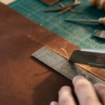 An artisan's hands cutting a thick piece of brown leather using a utility knife and a metal ruler on a green cutting mat. In the background, other leatherworking tools like a wooden-handled awl and a stitching punch rest on the workbench, surrounded by scattered leather scraps. The focus is sharp on the blade making the precise cut along the edge of the ruler.