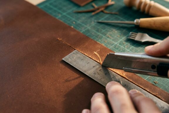 An artisan's hands cutting a thick piece of brown leather using a utility knife and a metal ruler on a green cutting mat. In the background, other leatherworking tools like a wooden-handled awl and a stitching punch rest on the workbench, surrounded by scattered leather scraps. The focus is sharp on the blade making the precise cut along the edge of the ruler.
